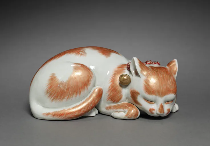 A white porcelain cat with orange-red patches curls asleep, a gold bell at its collar; photographed against a soft grey backdrop.