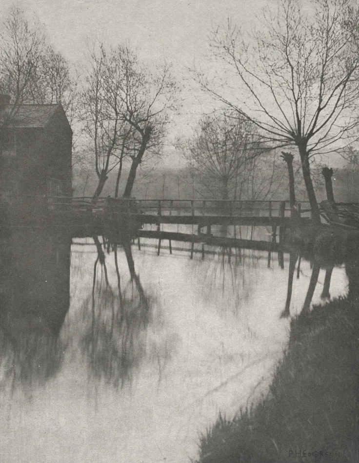 Willows and a wooden footbridge reflected in a calm stream beside a barn; hazy, low-contrast grey morning light.