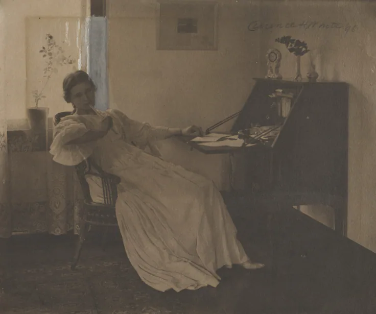 A woman in a white dress leans back from a writing desk, pen in hand, in a sparse room with flowers and a framed print; pencil signature at top.