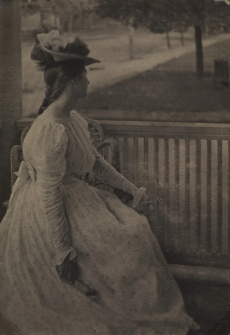 A woman in a pale dress and plumed hat sits on a porch rail looking out at a tree-lined street; soft summer light.