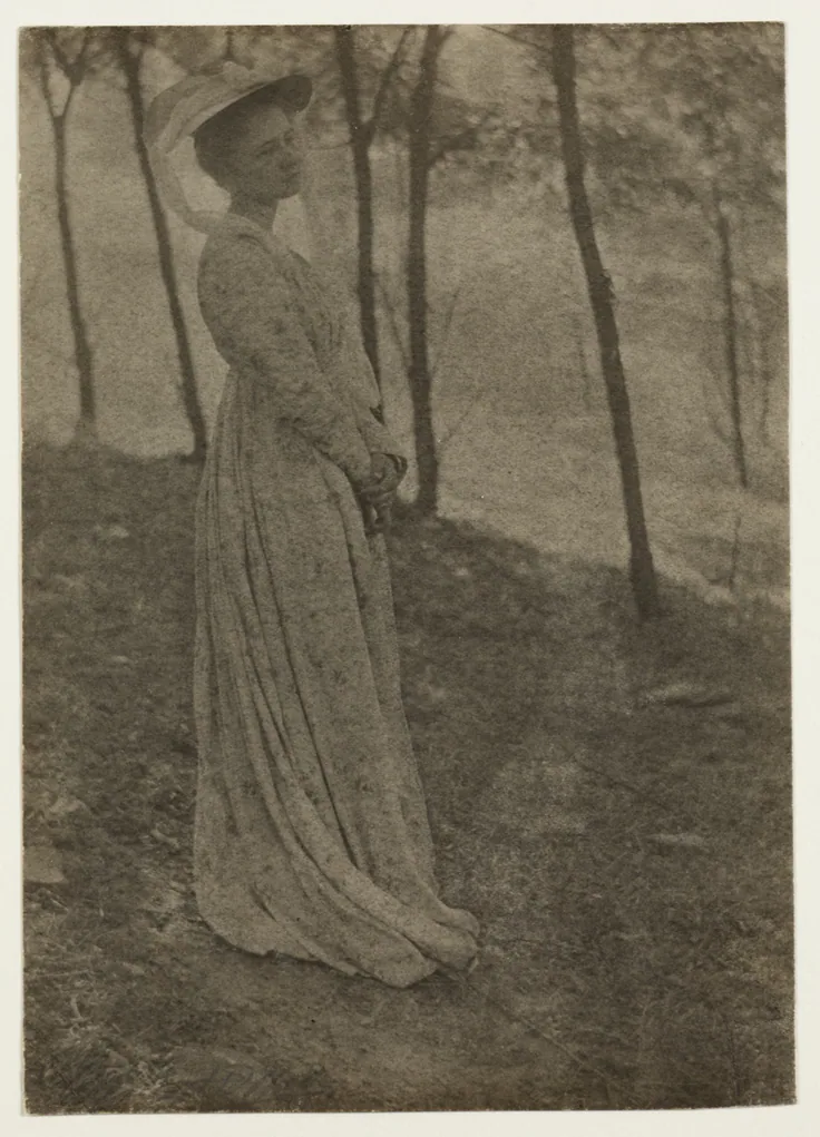 A woman in a long patterned dress and hat stands among slender trees on a slope; soft-focus platinum tones.