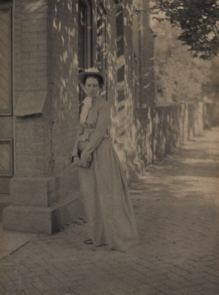 A woman in a long dress and boater hat stands by a stone building on a brick sidewalk, dappled with leaf shadows; soft olive tones.