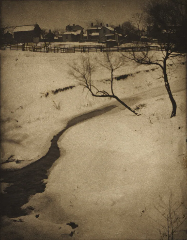 A dark stream winds through a snowy field toward bare trees and farmhouses on the ridge; soft platinum tones.