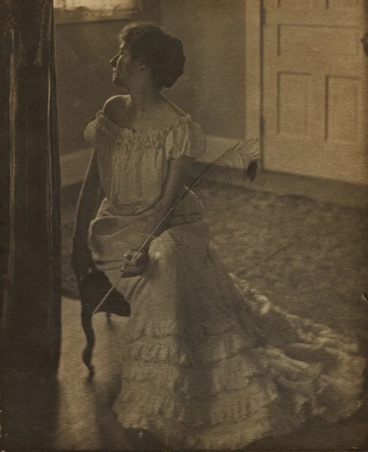 A woman in a ruffled white gown sits by a sunlit doorway holding a long peacock feather; quiet domestic light in warm platinum greys.