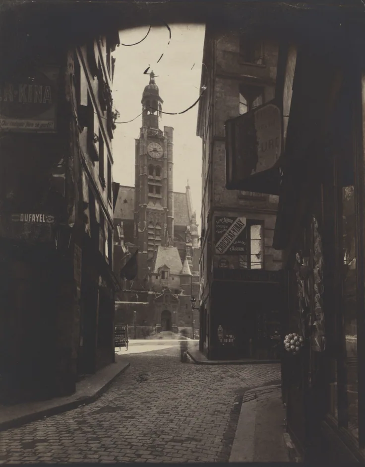 A narrow cobbled Paris street opens toward a church tower, dark shopfronts framing a bright patch of light.