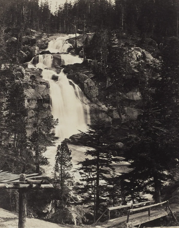A cascading torrent tumbles through dark pines and boulders toward a timber footbridge; crisp black and white with bright water.