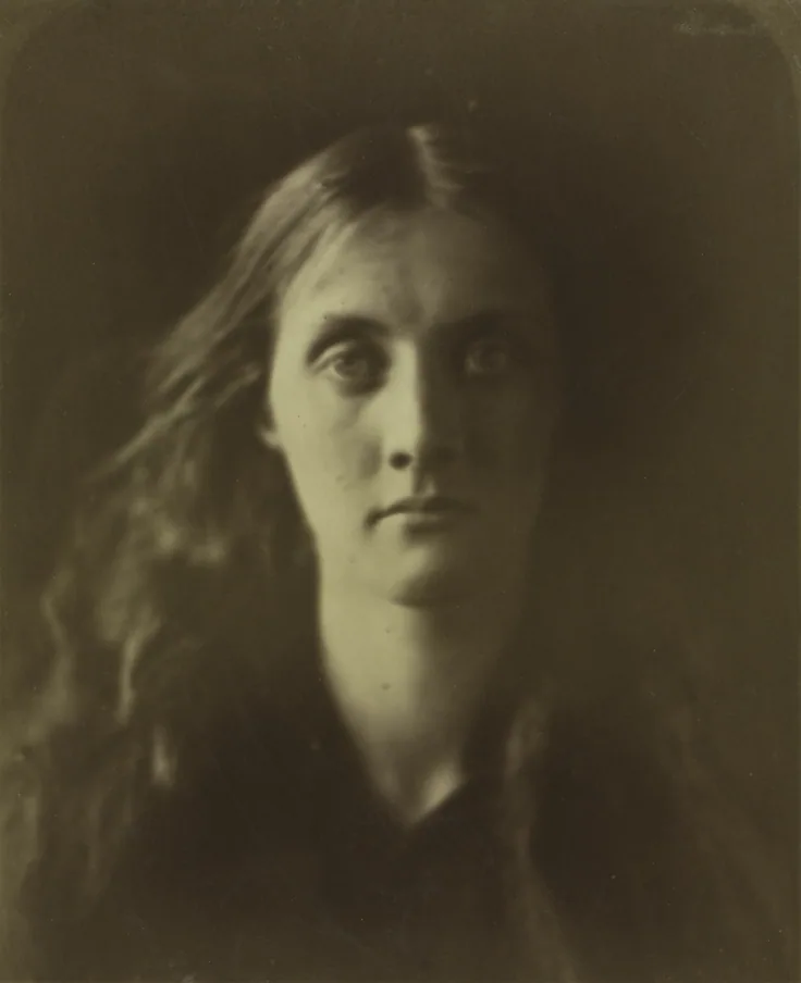 Close, soft-focus portrait of a young woman with long loose hair looking straight at the camera; her face glows against deep olive-brown shadow.