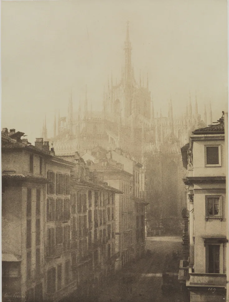 The spires of Milan Cathedral loom faintly through haze at the end of a canyon of tall buildings; pale ivory sepia.