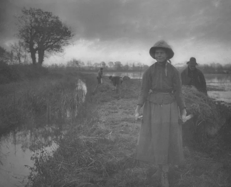 A young woman in a sunbonnet stands on a marsh path while men load hay behind her; a dark tree and a wide grey sky.