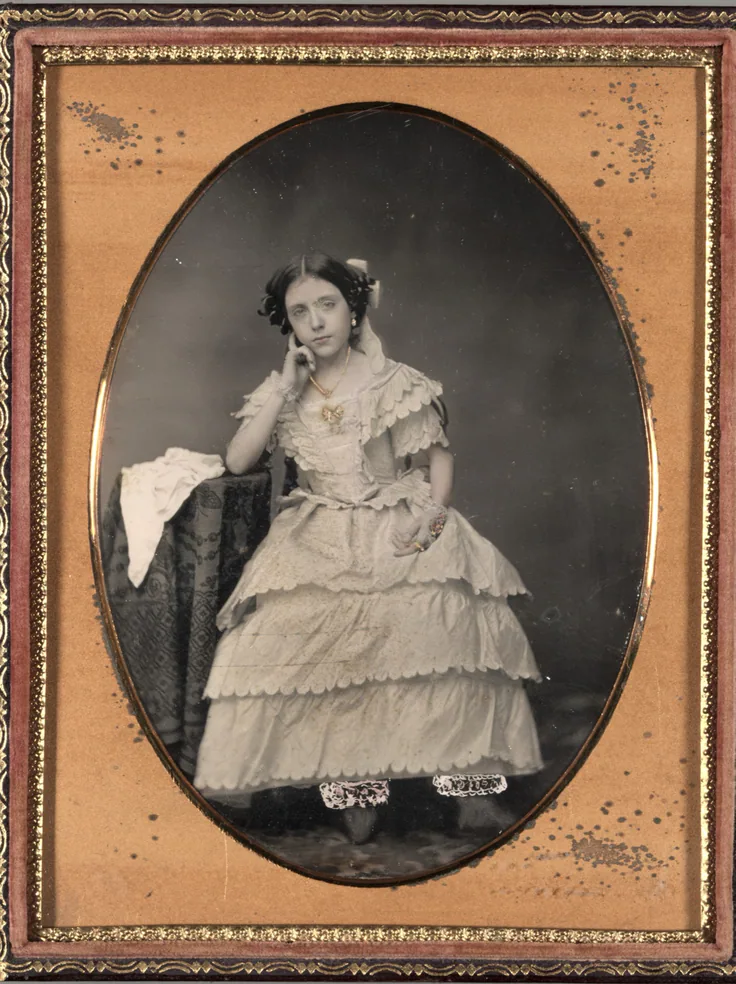 A young woman in a tiered ball dress poses by a draped table, framed in a gilt oval mat; a softly tinted daguerreotype.
