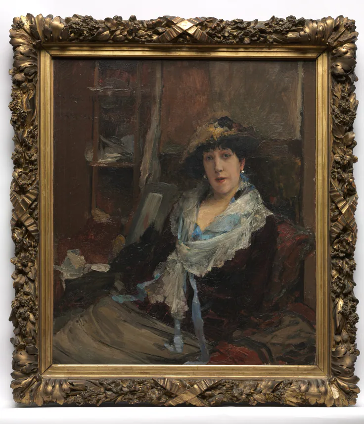 An actress in a dark dress, lace shawl and plumed hat sits in a dim studio, shown in its ornate gilt frame.