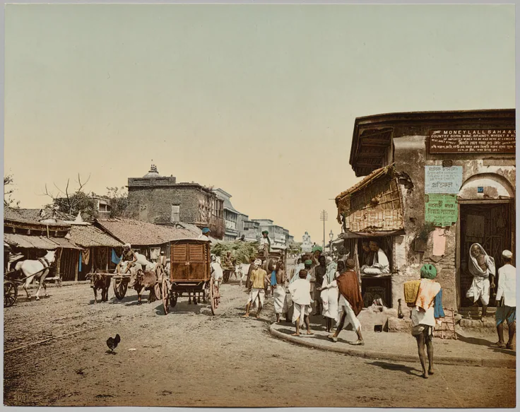 A busy dusty street with carts, a horse-drawn carriage, shops and people in white, in soft photochrom color.