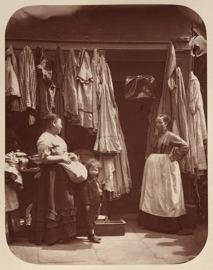 Two women and a boy stand in front of a London shop hung with dresses and coats; warm reddish-brown woodburytype.
